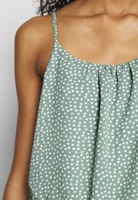 Top sans manches vert avec des pois blancs, présentant un col froncé et de fines bretelles, fabriqué dans un tissu doux et texturé.