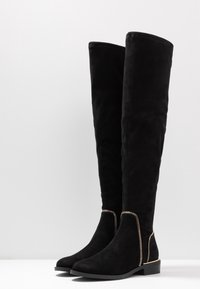 Knæhøje sorte ruskindsstøvler med en glat tekstur, der har gyldne lynlåsdetaljer langs siden og en flad sål.