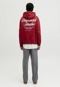 Rød hættetrøje med hvid tekst på ryggen. Kombineret med grå brede bukser og en hvid skjorte under.