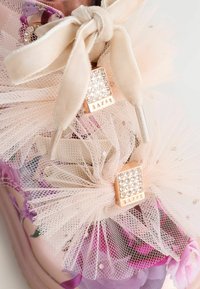 Vista ravvicinata di una scarpa con motivo floreale, dotata di un fiocco in tessuto color crema e fiori in tulle rosa decorativi con borchie di strass dorati, etichettata "BAXER."