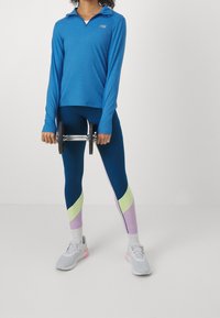 Osoba držící činku oběma rukama, oblečená v modrém tričku s dlouhým rukávem, kalhotách se dvěma barvami, bílých ponožkách a šedých teniskách.