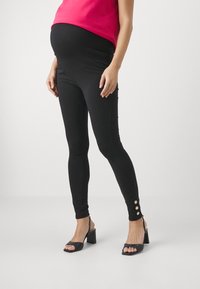 Svart gravide leggings med bred linning, elastisk stoff, figursydde design og gullknapper ved anklene.