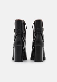 Botas de tobillo de cuero negro con un acabado texturizado, que cuentan con un tacón ancho y cuadrado, cremalleras laterales y un acento de correa en la parte trasera.