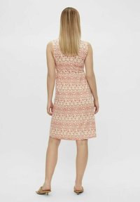 Mujer con cabello rubio lacio, vistiendo un vestido de tirantes de longitud hasta la rodilla en tonos rosa y beige con un patrón, y sandalias de tacón bajo beige, de pie de espaldas.