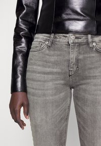 Top de manga larga de cuero negro combinado con jeans de mezclilla grises. Los jeans presentan un ligero desvanecimiento, bolsillos frontales y un cierre con botón plateado.