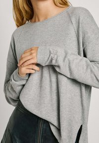 Pull gris clair à coupe décontractée, manches raglan et petites fentes latérales. Poignets et encolure côtelés ; texture douce. Jupe en cuir noir en dessous.