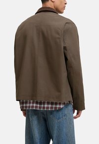 Chaqueta marrón de algodón con cuello de canalé, corte recto y dos bolsillos laterales, combinada con una camisa de cuadros y unos vaqueros holgados.