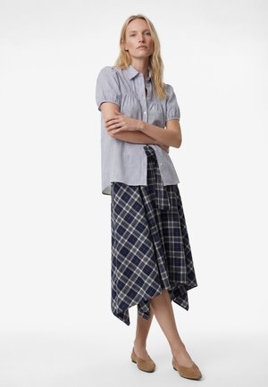 Vaaleatukkainen nainen seisoo käsivarret ristissä yllään lyhythihainen ruudullinen paitapusero, tummansininen ruudullinen epäsymmetrinen hame ja vaaleanruskeat tasapohjaiset kengät valkoisella taustalla.