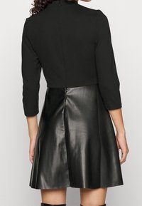 Robe noire avec un haut ajusté et une jupe en cuir évasée. Manches longues, col montant, texture lisse et fermeture éclair au dos.