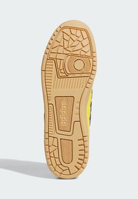 Semelle en caoutchouc beige d'une basket Adidas avec des motifs de semelle géométriques et en zigzag, montrant le nom de la marque embossé près du talon.