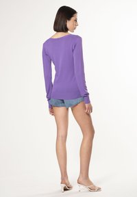 Suéter morado de manga larga con puños y dobladillo acanalados, combinado con pantalones cortos de mezclilla. Modelo vistiendo zapatos de tacón blancos, mostrado desde la parte de atrás.