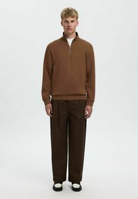 Brauner Zip-Pullover mit hohem Kragen, kombiniert mit locker sitzenden dunkelbraunen Hosen und schwarzen Schuhen mit weißen Akzenten.