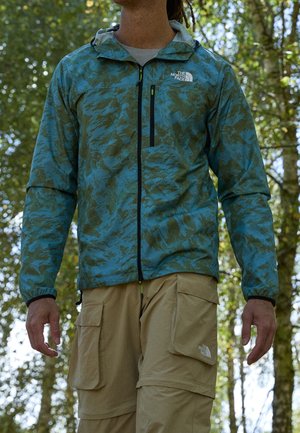 Mężczyzna noszący niebiesko-zieloną wzorzystą kurtkę i beżowe spodnie bojówki na zewnątrz, w tle drzewa.