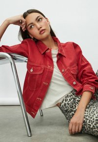 Red denim jacket with front pockets, metal buttons, and contrast stitching, paired with a white shirt and leopard print jeans. Smooth texture.