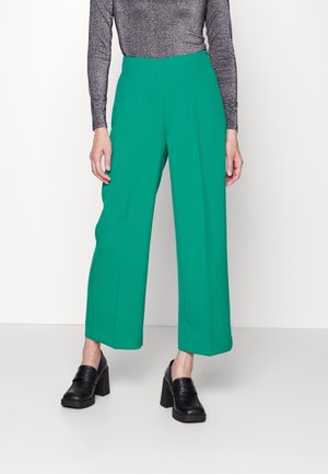 Kobieta ubrana w zielone spodnie o wysokim staniku i szerokich nogawkach, czarne loafersy na masywnym obcasie oraz dopasowaną, metalicznie szarą bluzkę z długim rękawem, stojąca na tle białej ściany.