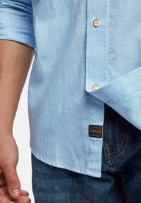 Hellblaues Hemd mit Knöpfen aus strukturiertem Stoff, mit orangefarbenen Akzenten an den Knöpfen und einem Markenschild am Saum. Getragen mit dunklen Jeans.