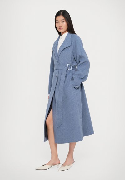 Femme portant un long manteau bleu ceinturé sur une chemise blanche et des mules crème à talons pointus, se tenant contre un fond blanc uni.