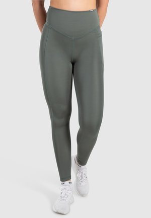 Hoch-taillierte, olivgrüne Leggings mit glatter Textur, die überkonturierte Nähte und Seitentaschen verfügt. Kombiniert mit weißen Sportschuhen.