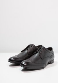 Chaussures habillées en cuir noir avec design à bout golf et détails brogues, bout rond et petit talon. Texture lisse et style classique à lacets.