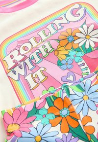 Puuvillapaita, jossa on monivärinen kukkakuva pinkissä, oranssissa ja sinisessä, ja se sisältää tekstin "ROLLING WITH IT" lihavoidulla fontilla.