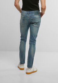 Šviesiai mėlyni džinsai, turintys siaurą kirpimą, šiek tiek blukimo ir dvi galines kišenes su užsegimais. Juosmens juosta turi etikėtę su prekės ženklo informacija.