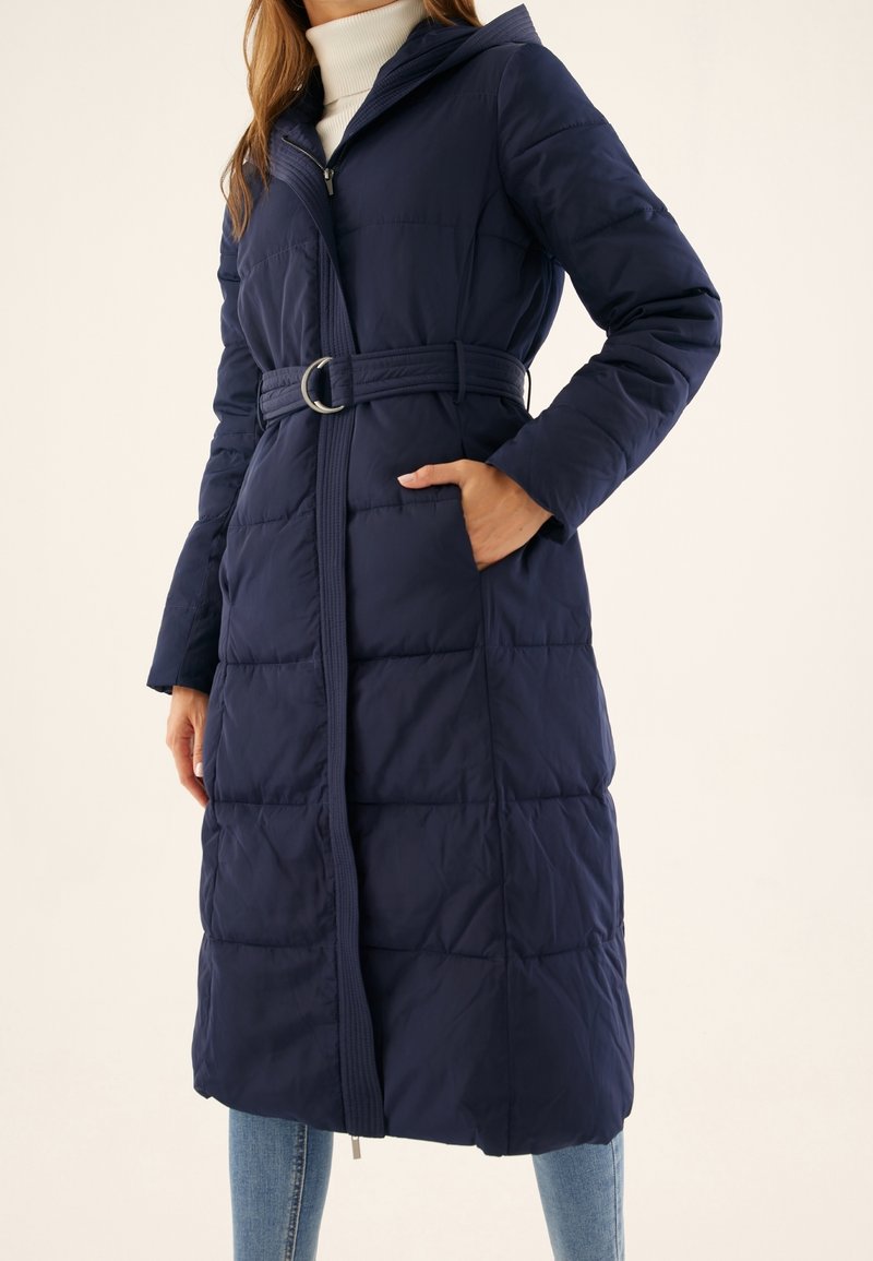 Femme portant un long manteau matelassé bleu marine avec une ceinture, main dans la poche, col roulé blanc et jeans bleu clair sur un fond uni.