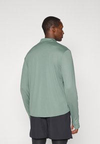Langarm-Poloshirt aus leicht grünem Stoff mit glatter Textur. Verfügt über einen klassischen Kragen und einen leicht verlängerten hinteren Saum.