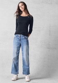 Schwarzes Langarmhemd kombiniert mit hellblauen weit geschnittenen Jeans. Die Schuhe sind weiße Sneakers. Einfaches, figurbetontes Design mit lässigem Look.