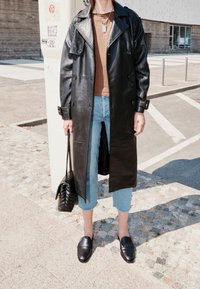 Abrigo largo negro de cuero tipo gabardina con diseño de doble botonadura, combinado con vaqueros crop de color azul claro y mocasines negros. Lleva un bolso negro acolchado.