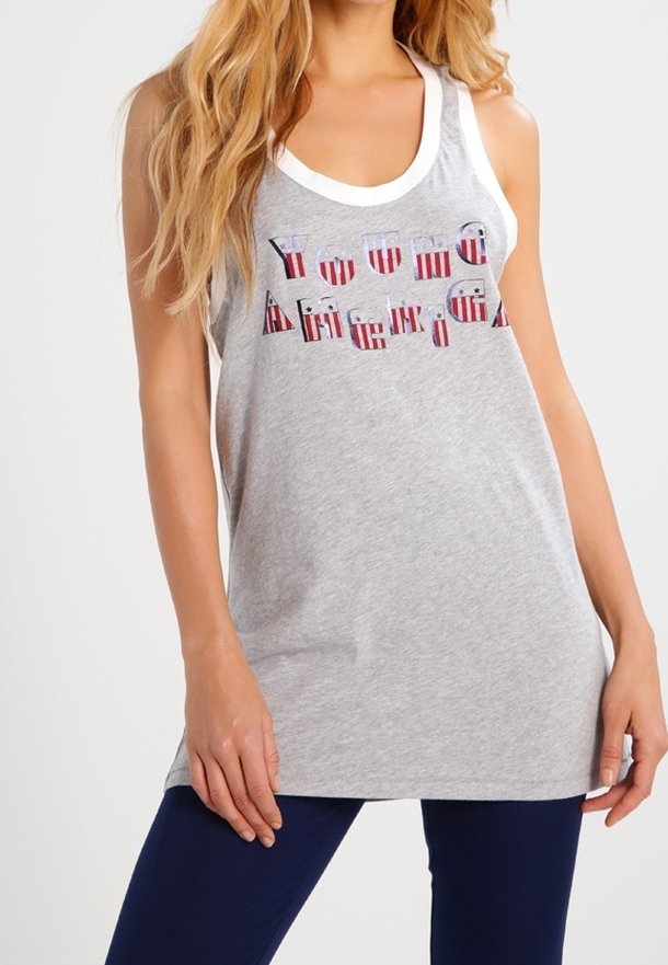 Femme portant un débardeur gris sans manches avec le texte « Young America » en lettres aux motifs rouge, blanc et bleu, assorti à un pantalon foncé.