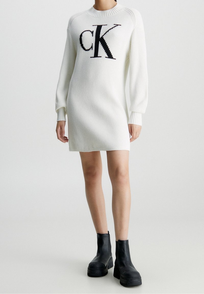 Abito maglione bianco a coste con il logo "CK" nero al centro, scollo rotondo e maniche lunghe leggermente a sbuffo. Stivali neri alla caviglia.