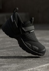 Chaussure de sport noire avec une tige texturée, des motifs matelassés, une large bride ornée d'accents argentés et une semelle arrondie pour le confort.