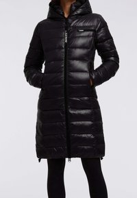 Cappotto lungo in piumino nero, trapuntato, con cappuccio, chiusura zip e tasche laterali indossato con leggings neri, mani nelle tasche.