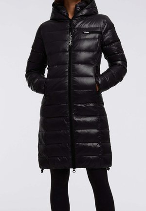 Manteau matelassé noir long avec capuche, fermeture éclair à l'avant et poches latérales, porté avec des leggings noirs, les mains dans les poches.