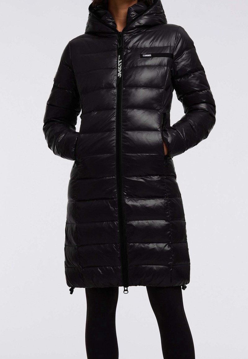 Cappotto lungo in piumino nero, trapuntato, con cappuccio, chiusura zip e tasche laterali indossato con leggings neri, mani nelle tasche.