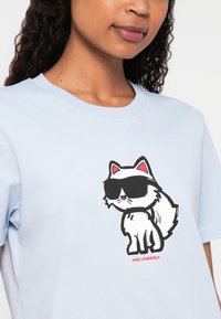 Světle modré tričko s potiskem karikatury bílého kočka s růžovými akcenty a slunečními brýlemi, tištěným černou barvou. Text "KARL LAGERFELD" v červené barvě ve spodní části.