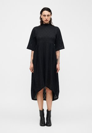 Person står vendt fremad iført en sort high-low kjole med albuelange ærmer og sorte ankelstøvler mod en enkel, lys baggrund.