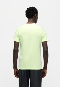 Ανοιχτό πράσινο βαμβακερό μπλουζάκι με κοντά μανίκια, στρογγυλή λαιμόκοψη και ομαλή υφή. Φοριέται με μαύρο σορτς, και φαίνεται από πίσω.