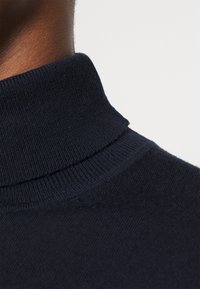 Dunkelblauer, gestrickter Pullover mit hohem Kragen und geripptem Textur, der eine glatte Oberfläche und ein eng anliegendes Kragendesign aufweist.