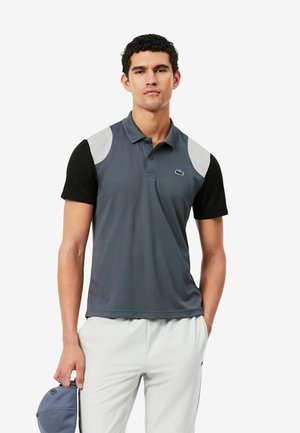 Homme portant un polo gris et noir avec des touches blanches, pantalon blanc, tenant une casquette grise dans sa main gauche sur un fond blanc.