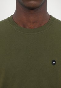 Tricou verde din bumbac, cu guler rotund și mâneci scurte. Prezintă un mic logo hexagonal negru cu o stea albă în zona stânga a pieptului.