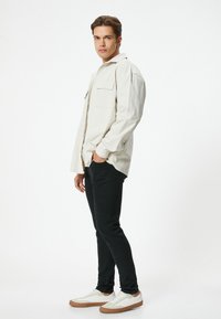 Camisa abotonada en color blanco roto para hombre con dos bolsillos en el pecho, pantalones negros ajustados y zapatillas blancas con suelas en tono marrón claro, presentados sobre un fondo liso.