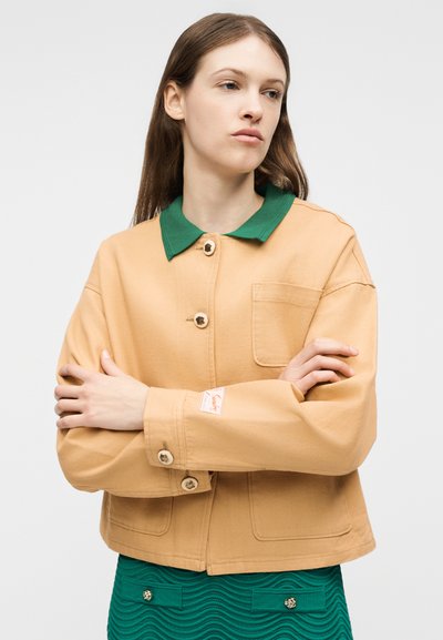 Νεαρή γυναίκα με σταυρωμένα χέρια, φορώντας μπεζ σακάκι με κουμπιά και πράσινο γιακά, καθώς και υφασμάτινη πράσινη φούστα, σε μονόχρωμο φόντο.