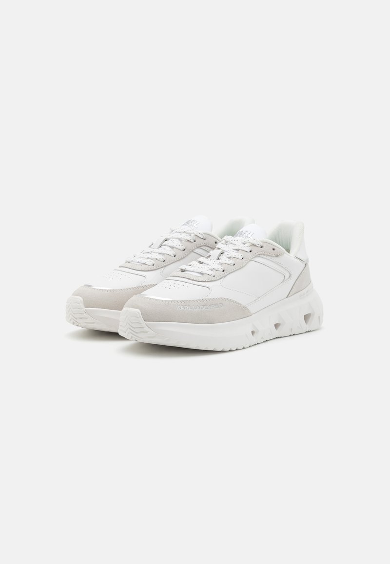 Zapatillas blancas con un frente de ante texturizado y paneles de cuero liso. Presentan cordones deportivos, una suela gruesa y un logotipo grabado en el lateral.