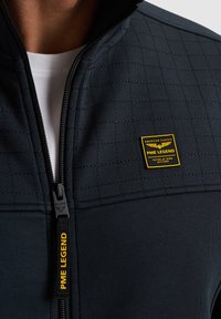 Schwarze Zip-Jacke aus strukturiertem Stoff mit einem Gitternmuster. Verfügt über ein gelbes Markenlabel und eine kontrastierende weiße Innenschicht.