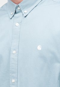 Ανοιχτό μπλε πουκάμισο από βαμβάκι με κλασικό γιακά, λευκά κουμπιά και μικρό κεντημένο λογότυπο στο στήθος.