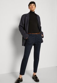 Marineblaue, taillierte Hose mit geradem Schnitt, kombiniert mit einem schwarzen Rollkragenpullover und einem langen dunklen Blazer. Schwarze Slipper vervollständigen den Look.