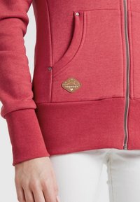 Rode zip-up hoodie gemaakt van zacht materiaal met ribbed manchetten en zoom. Bevat een zijzak en een bruine label met witte tekst.