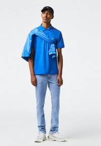 Blaues Poloshirt, hellblaue Jeans und weiße Sneaker. Eine blaue Jacke liegt lässig über den Schultern. Das Poloshirt trägt das Lacoste-Logo.