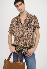 Camisa de manga corta con estampado de leopardo y un logo negro, combinada con pantalones cortos negros con patrón de texto y un bolso tote marrón brillante. Unos vaqueros azules completan el look.